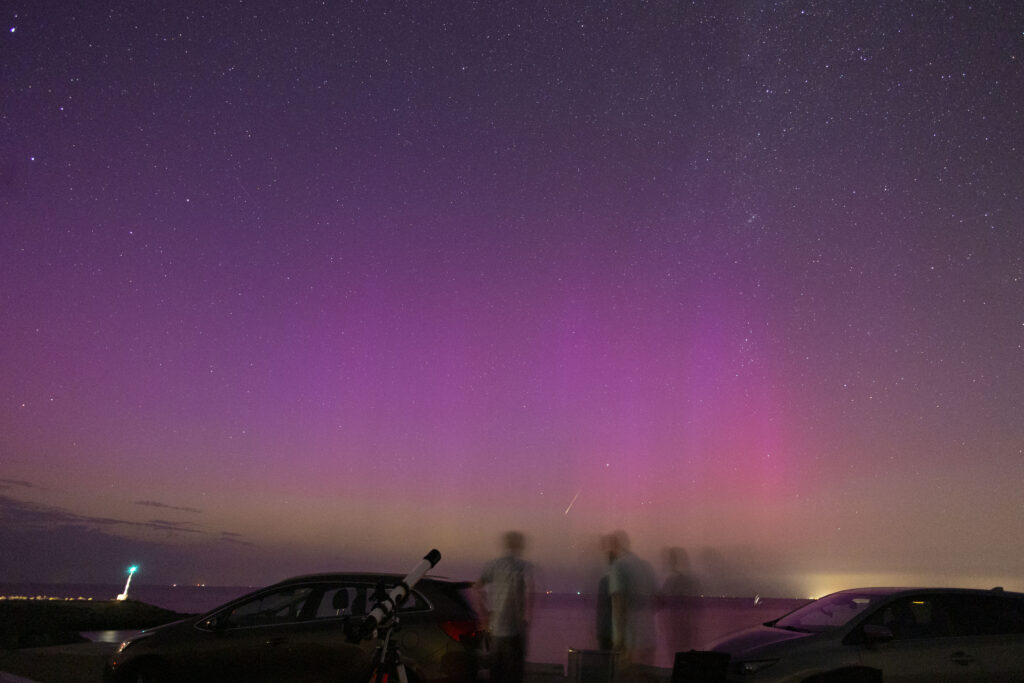 "Selfie" van 4 personen met poollicht en een heldere perseïde richting de horizon. De personen staan tussen auto's met een kleien klassieke telescoop erbij.